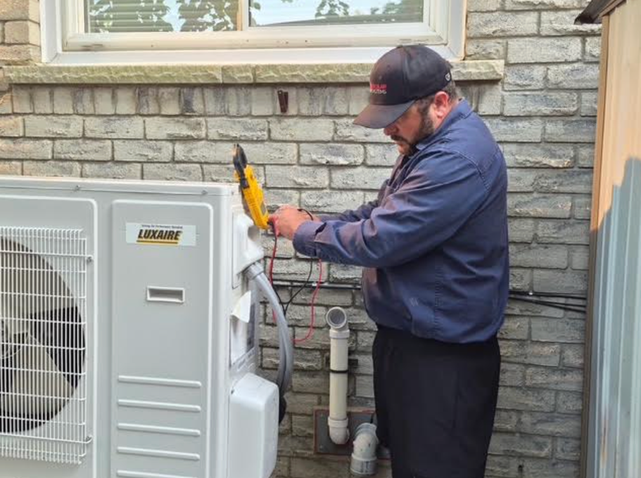 Technician using a yellow tool to service an outdoor HVAC unit beside a stone house