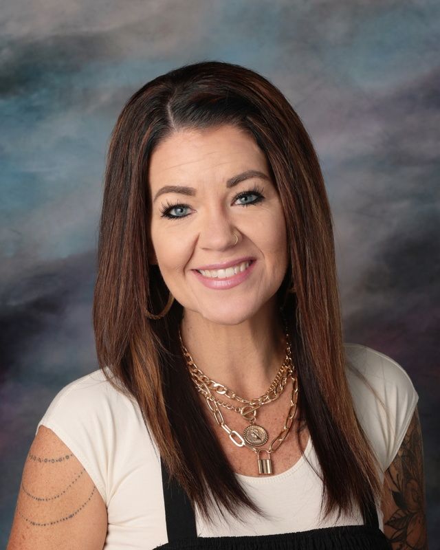 A woman wearing a yellow cardigan and a white shirt is smiling in front of a blue background.