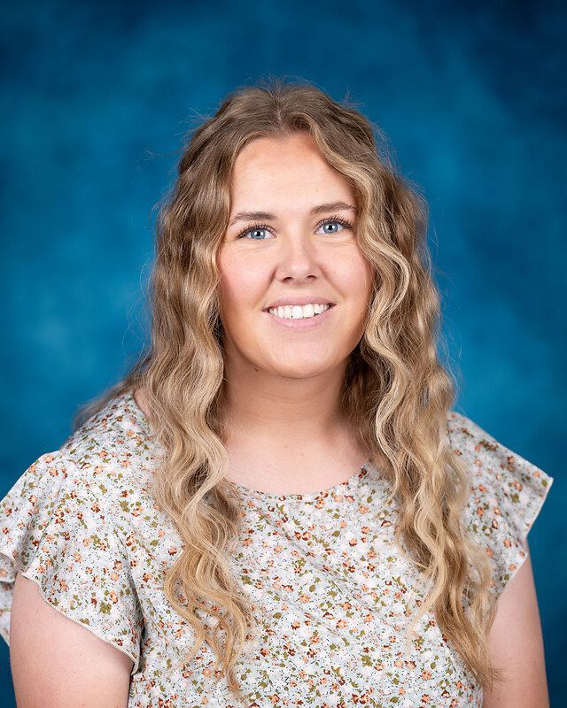 A young woman with long blonde hair is smiling for a picture in front of a blue background.