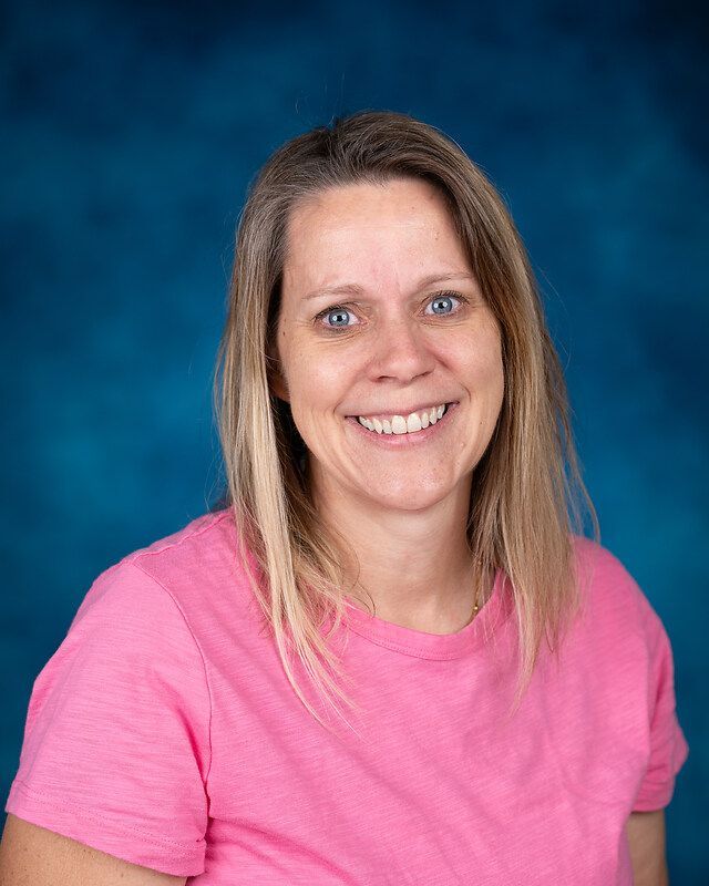A woman in a pink shirt is smiling in front of a blue background.