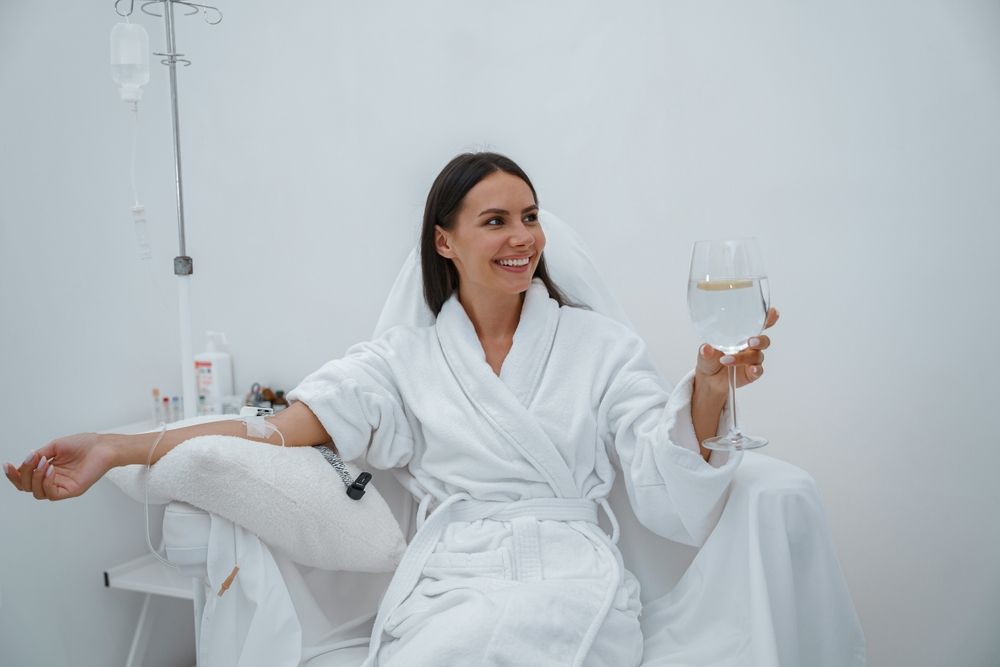 Woman in a white robe receiving an IV drip, smiling while holding a glass of clear liquid. White room.
