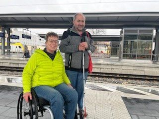 Eine Frau im Rollstuhl und ein Mann mit Langstock am Bahnsteig.