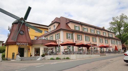 Hotel Gasthof Zur Windmühle.  Historisches Gebäude mit Windmühle. Davor Terrasse mit Sonnenschirmen.