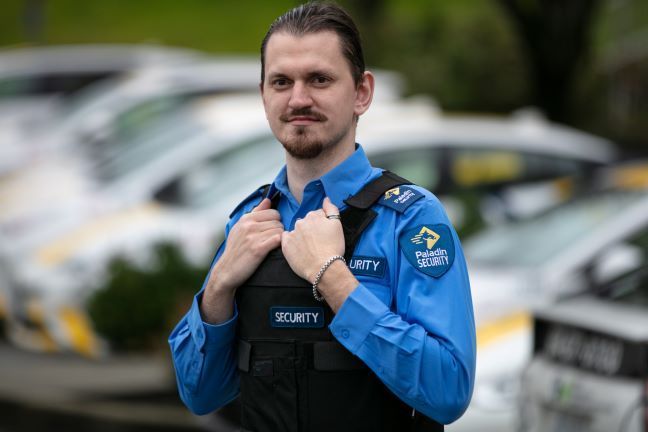 A man in a blue shirt and black vest is standing in front of a parking lot.