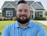 Man with beard smiling in front of a house; wearing a blue polo shirt.