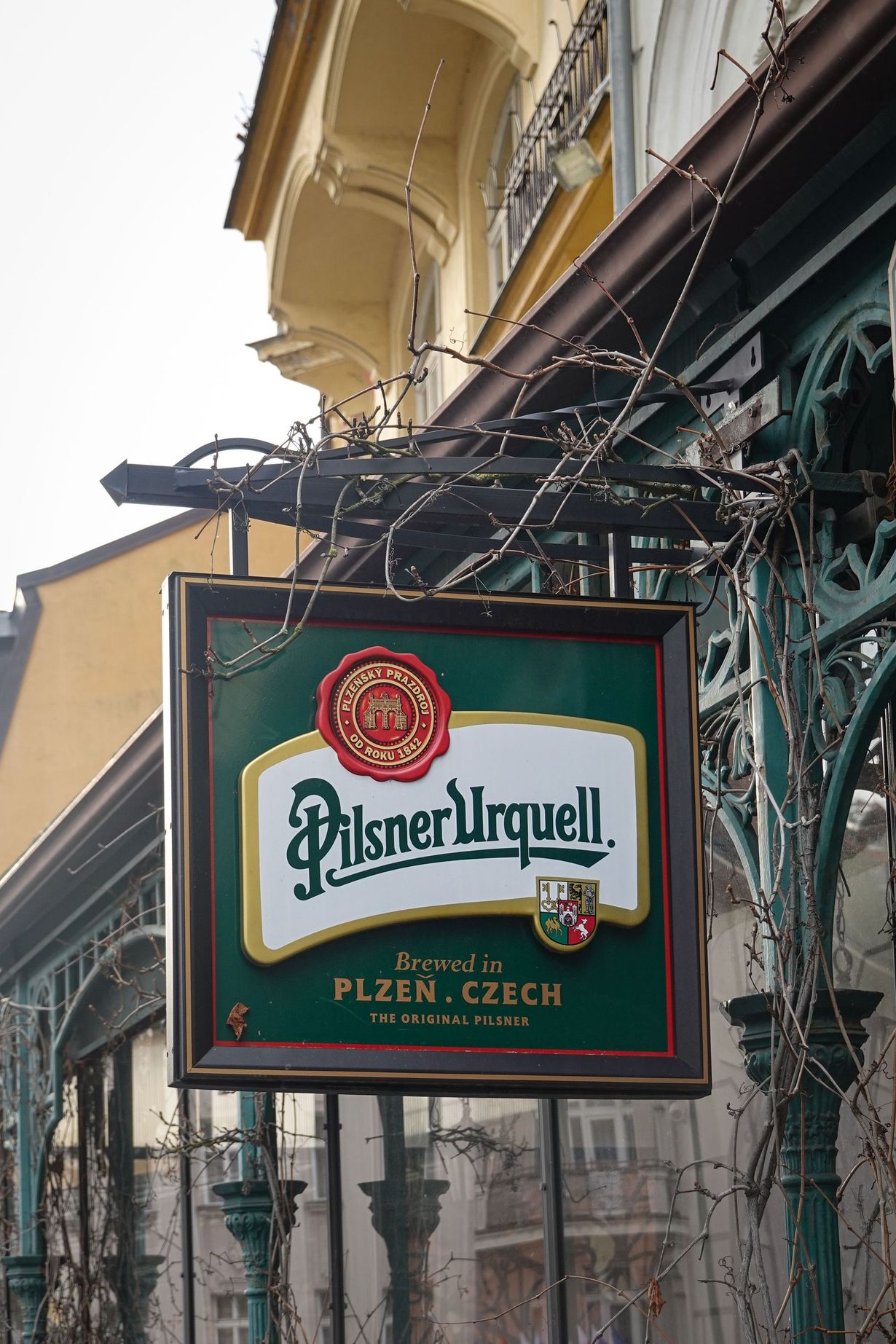 Insegna verde e rossa del pub Pilsner Urquell appesa a un edificio in una strada.