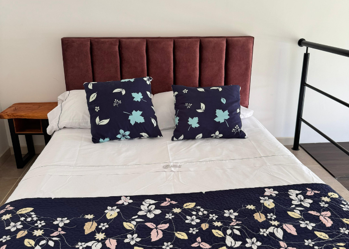 Dormitorio con cabecero color burdeos, ropa de cama blanca, almohadas estampadas y camino de mesa con flores.