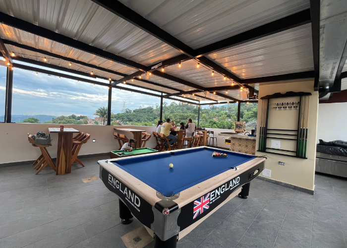 Área recreativa al aire libre con mesa de billar, mesas y gente. Ofrece vistas a la montaña y guirnaldas de luces.