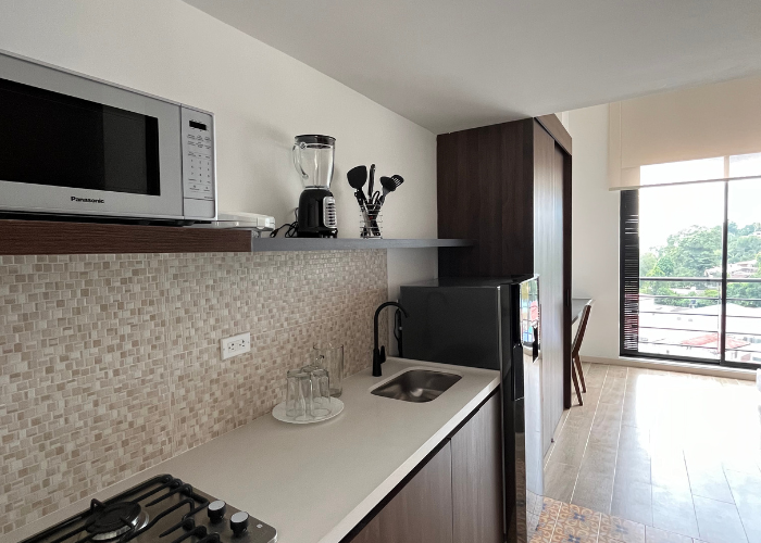 Cocina pequeña con microondas, licuadora, fregadero y nevera pequeña. Ventana a la derecha.