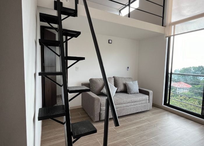 Interior con escalera de caracol, sofá gris, loft y gran ventanal con vistas.