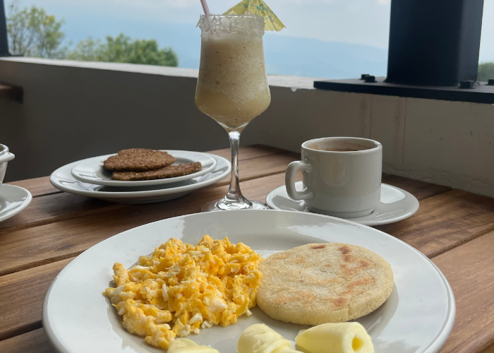 Plato de desayuno con huevos revueltos, arepa, plátano, galletas, café y cóctel en una mesa de madera.