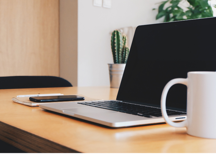 Computadora portátil, teléfono, taza y cactus sobre un escritorio de madera en un espacio de trabajo luminoso.