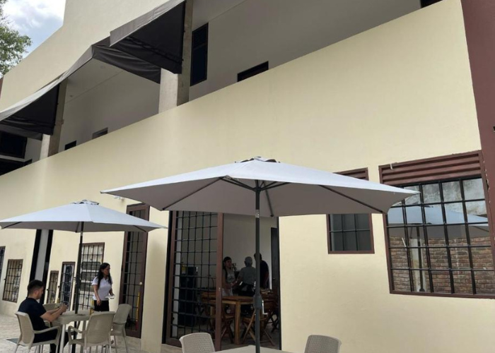 Cafetería al aire libre con edificio de color crema, sombrillas grises, mesas y gente sentada/de pie.