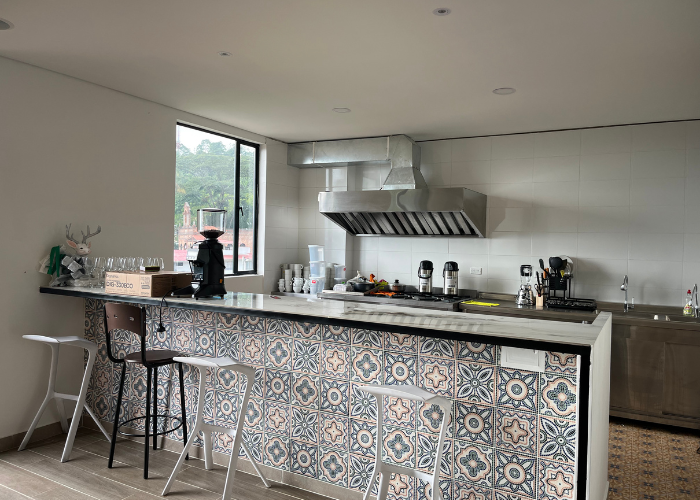 Barra de cocina con azulejos estampados, taburetes, equipamiento y una ventana con vista a la vegetación.