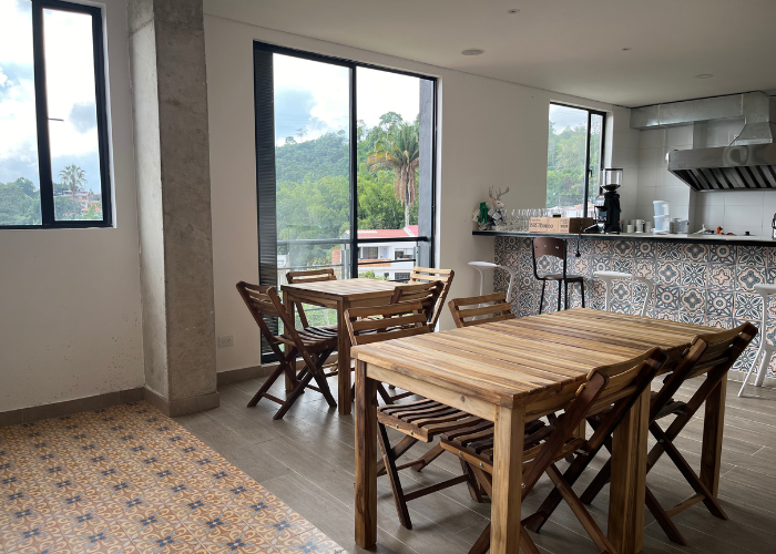 Interior de cafetería con mesas y sillas de madera cerca de grandes ventanales, con vistas a una ladera.