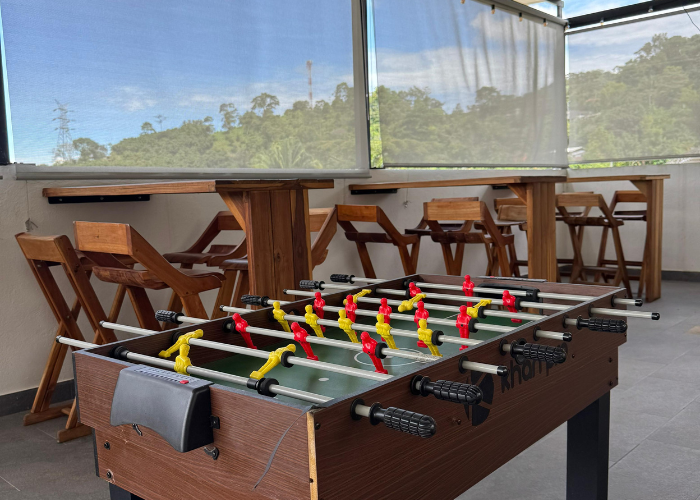 Mesa de futbolín en zona exterior con taburetes de madera y vistas a los árboles.