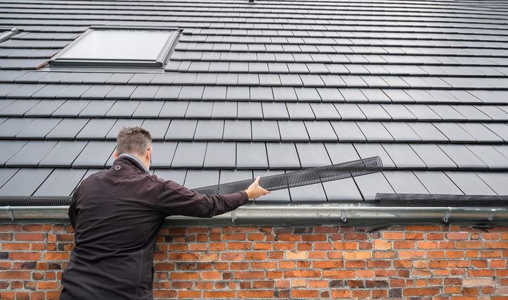 A person installs a black gutter guard strip along the edge of a dark-tiled roof above a brick wall.