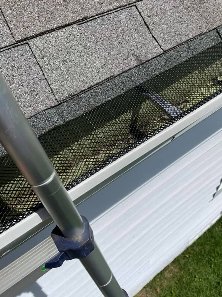 Gutter with black mesh guard, filled with debris, with a pole angled towards it. Gray shingles visible.