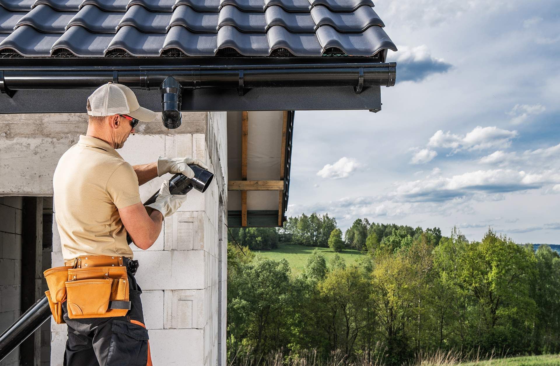 A construction worker in a tan shirt and tool belt installs a black gutter downspout on a house with a tile roof.