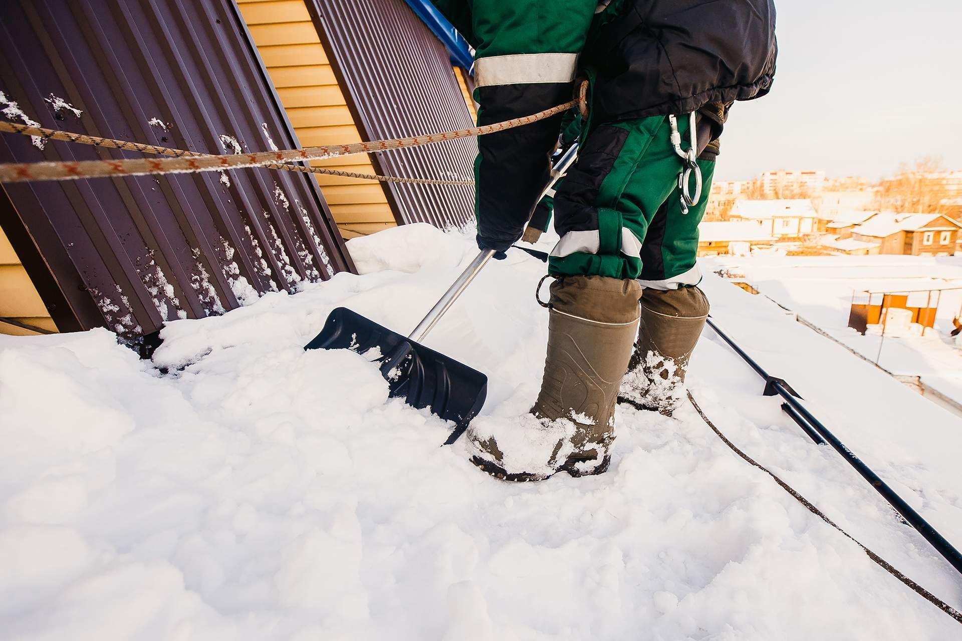Person shoveling snow from a roof, wearing green overalls, boots, and a safety harness.
