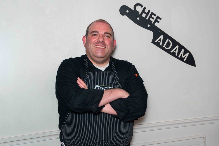 A man in an apron is standing in front of a wall that says chef adam