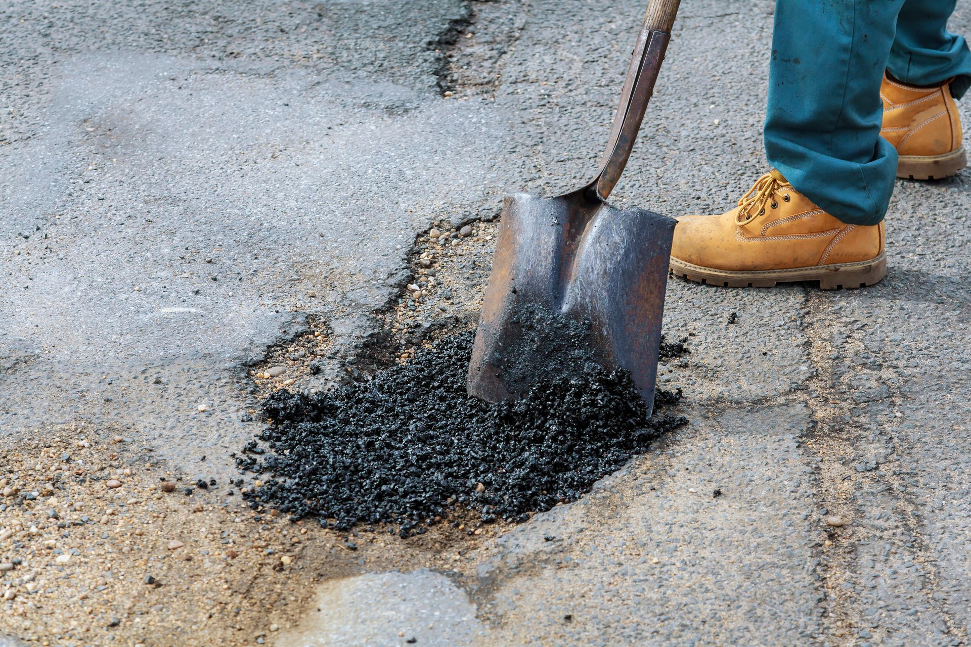 Shovel spreading black asphalt patch on cracked pavement beside a worker’s tan boots