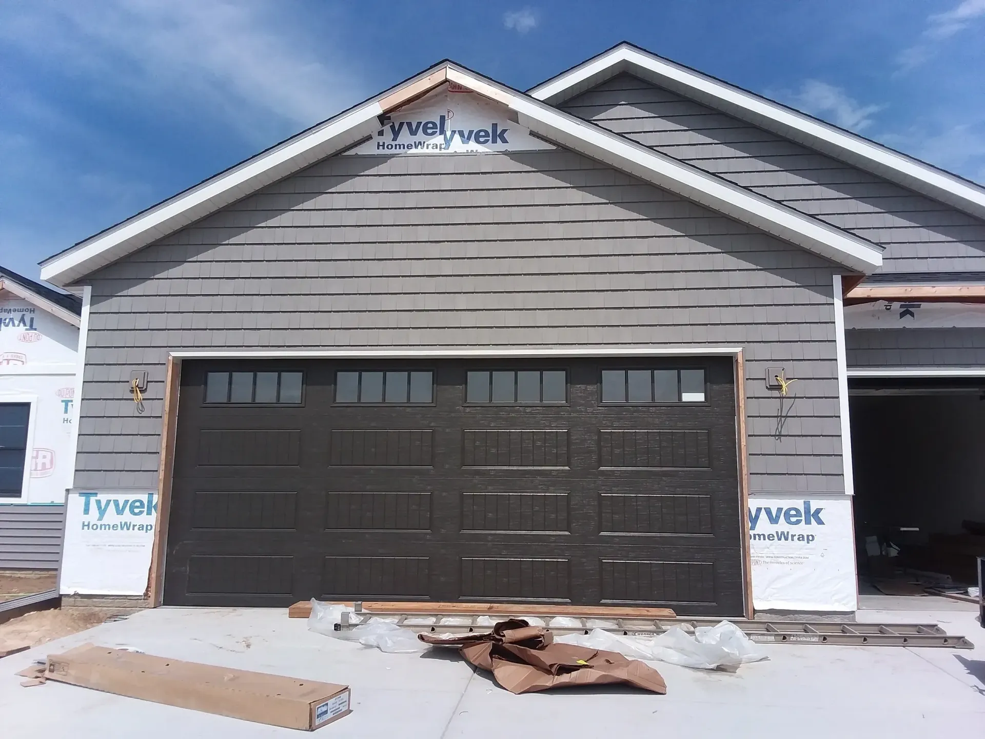 New construction garage with dark brown door and gray siding, blue sky visible.