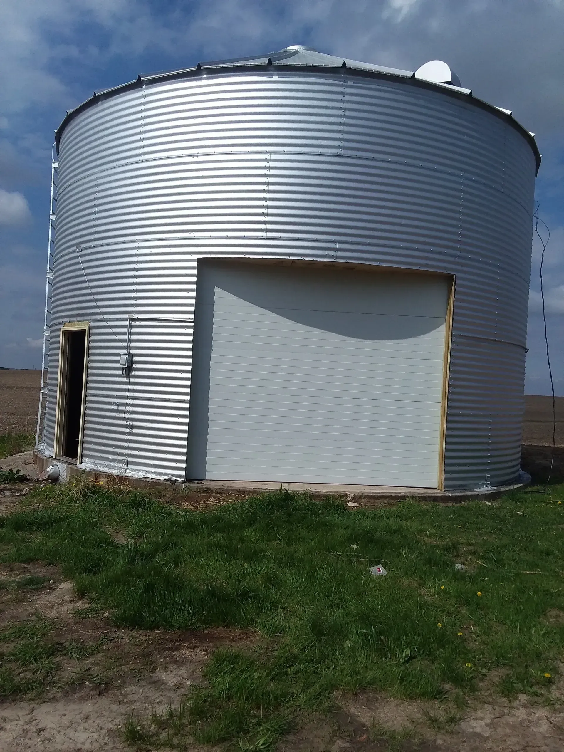 Silver grain silo with two doors in a field, blue sky background.