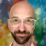 A bald man with a beard and glasses is smiling in front of a colorful background.