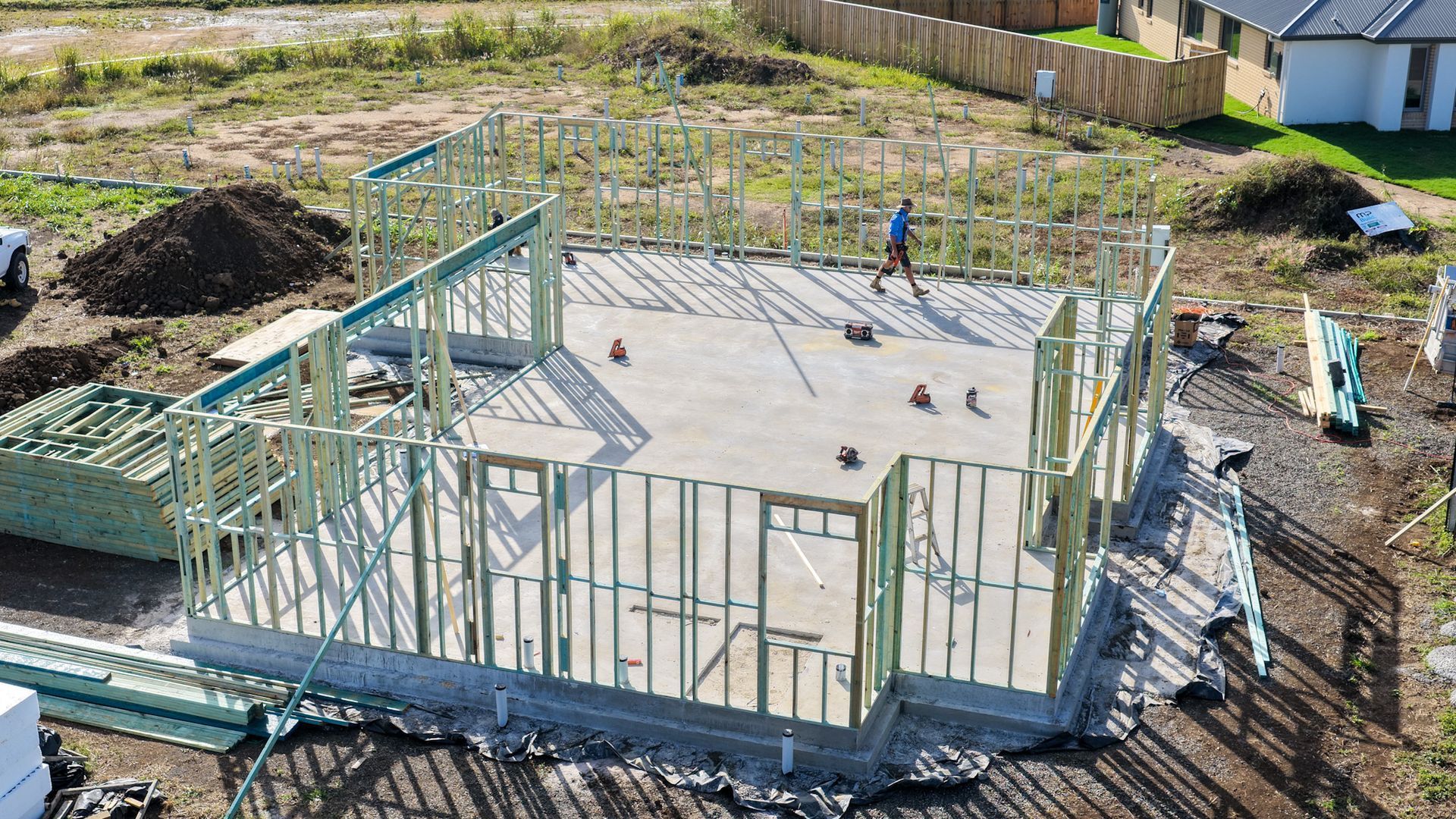 House foundation under construction with framed walls on a cleared lot, workers in the background