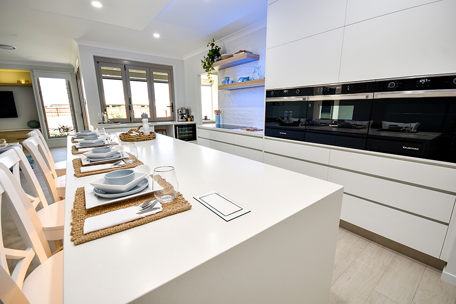 Bright modern white kitchen with large island, set dining placemats, and sleek black built-in appliances.