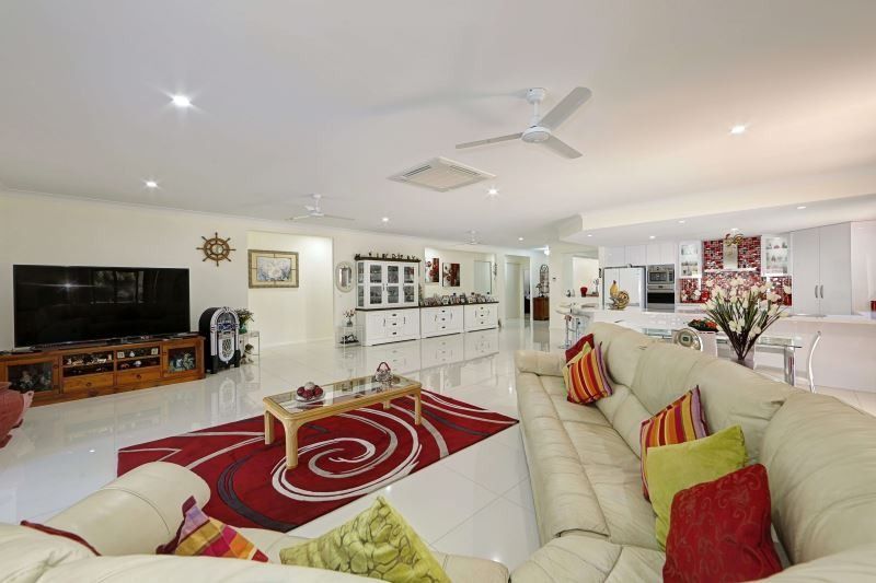 Bright open living room with white sofas, red rug, TV, ceiling fan, and a glimpse of the kitchen area.