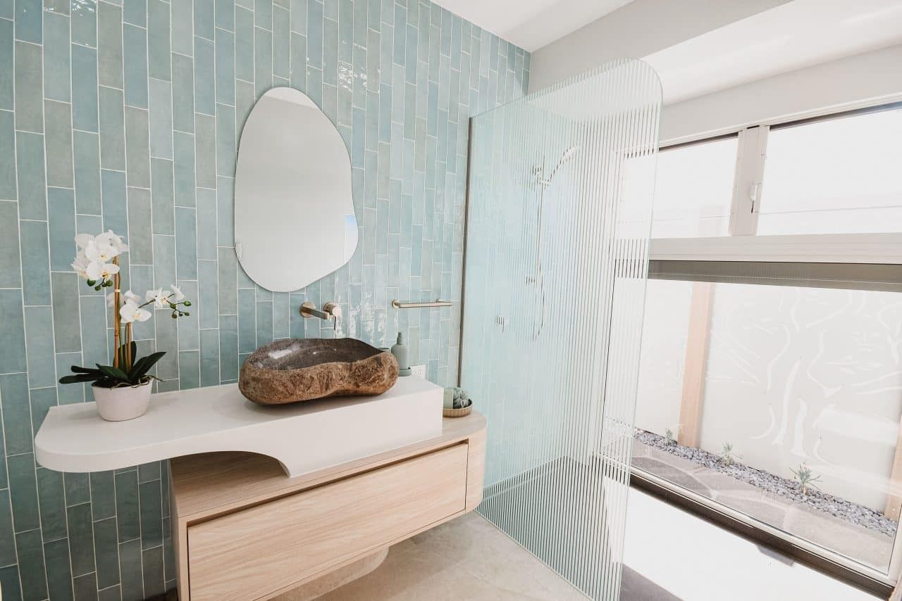 Bright modern bathroom vanity with blue tile wall, oval mirror, white sink, and sunlight through frosted window.