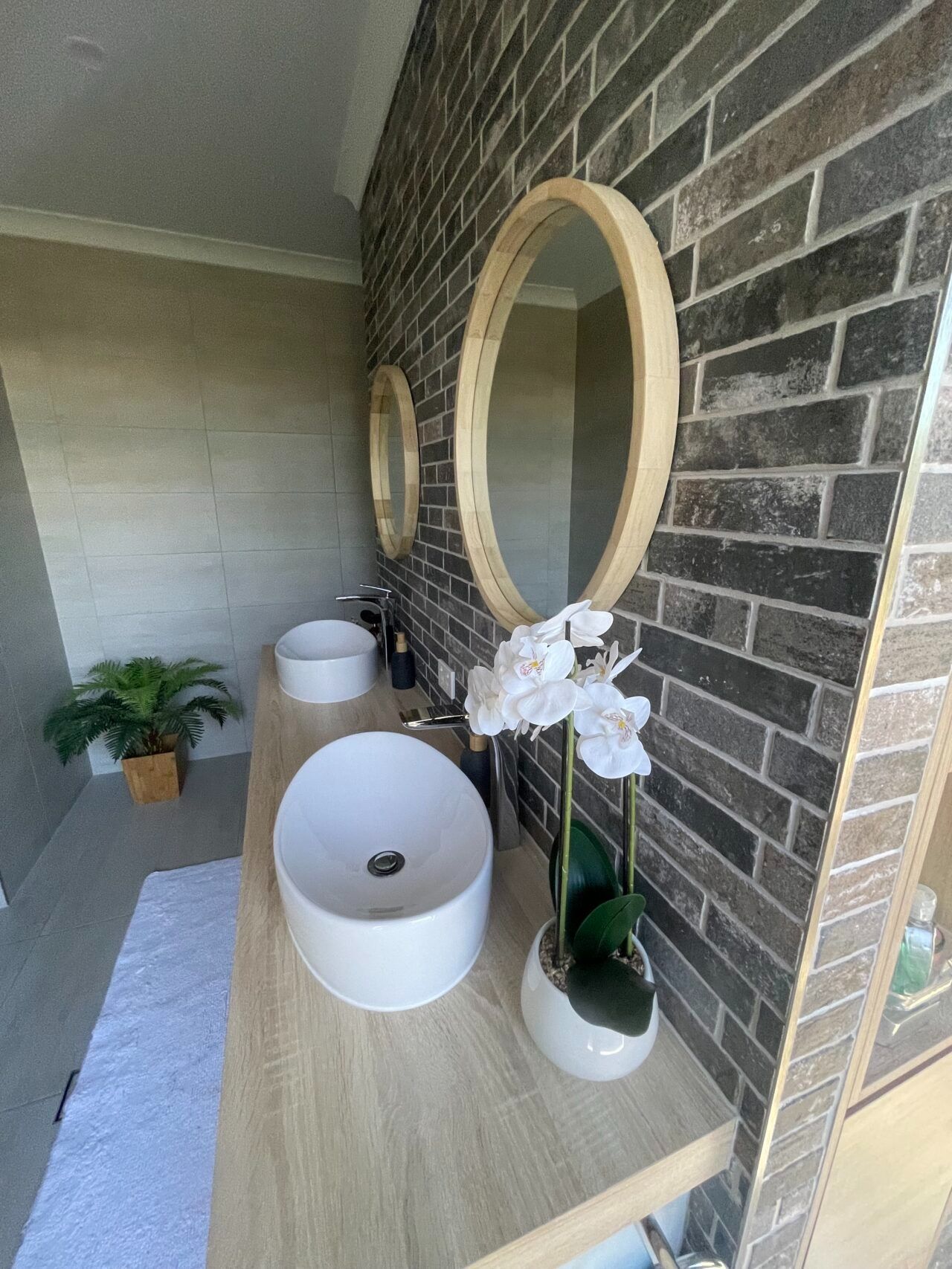 Modern bathroom vanity with round mirrors, white vessel sinks, and an orchid against a brick accent wall.