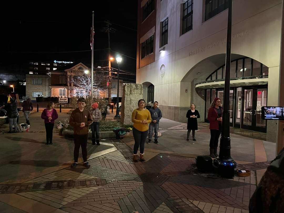 Group of people standing outdoors at night, possibly singing. City street with buildings and lights.