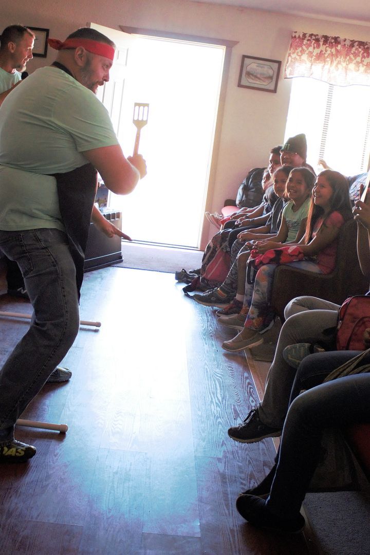 Man with spatula, wearing red bandanna, facing a seated group. Indoors, sunny, wooden floor.