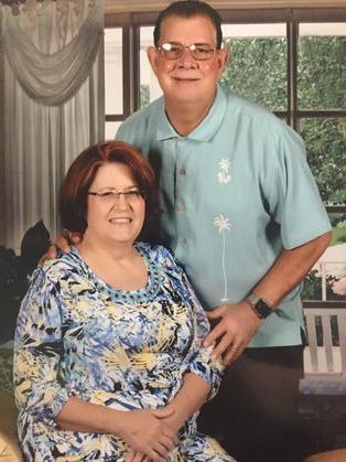 Couple poses for a photo. The woman sits in a patterned top, the man in a blue shirt. Behind a window.