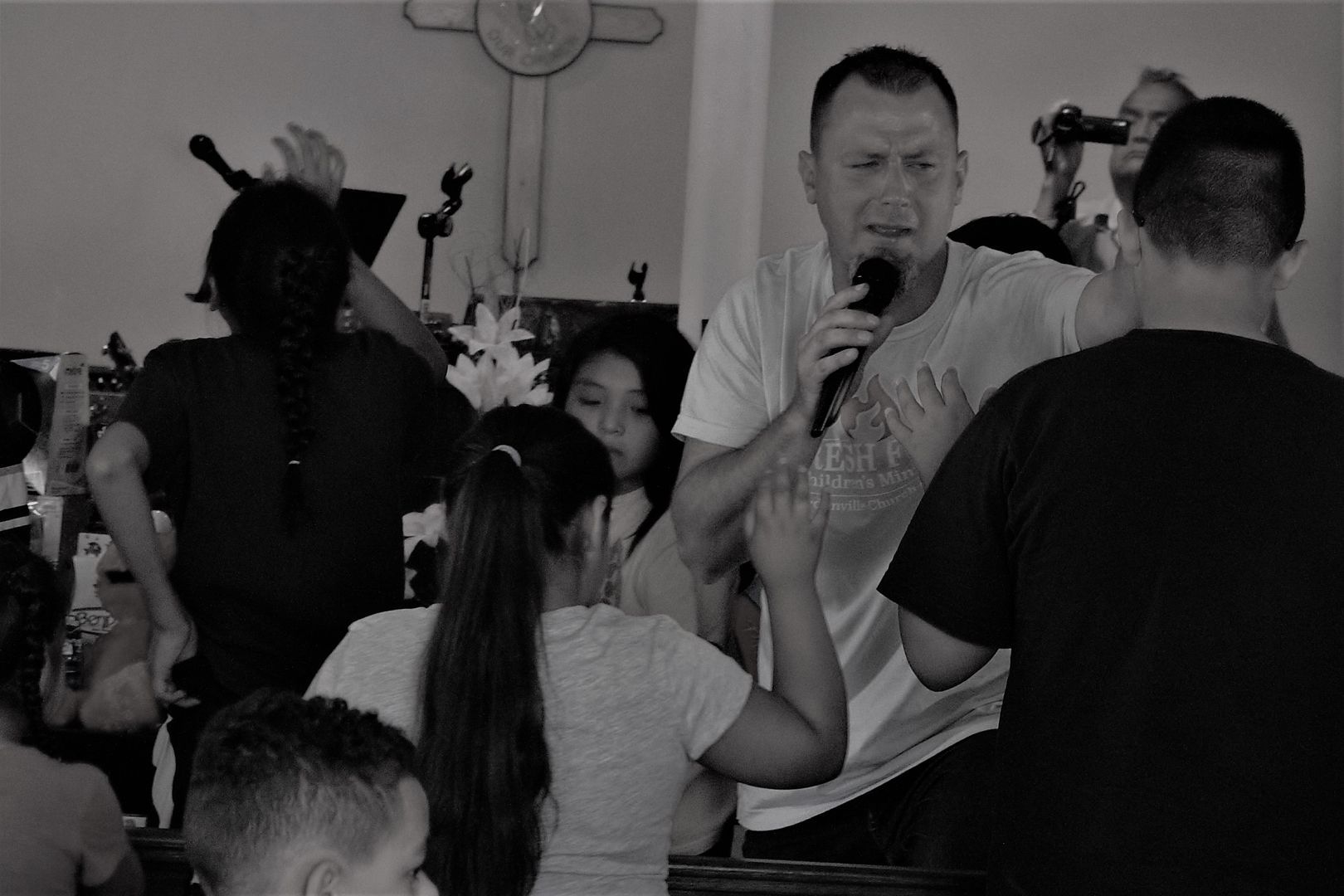 People in a room with a man speaking into a microphone. Others have their hands raised. Cross on the wall. Black and white.