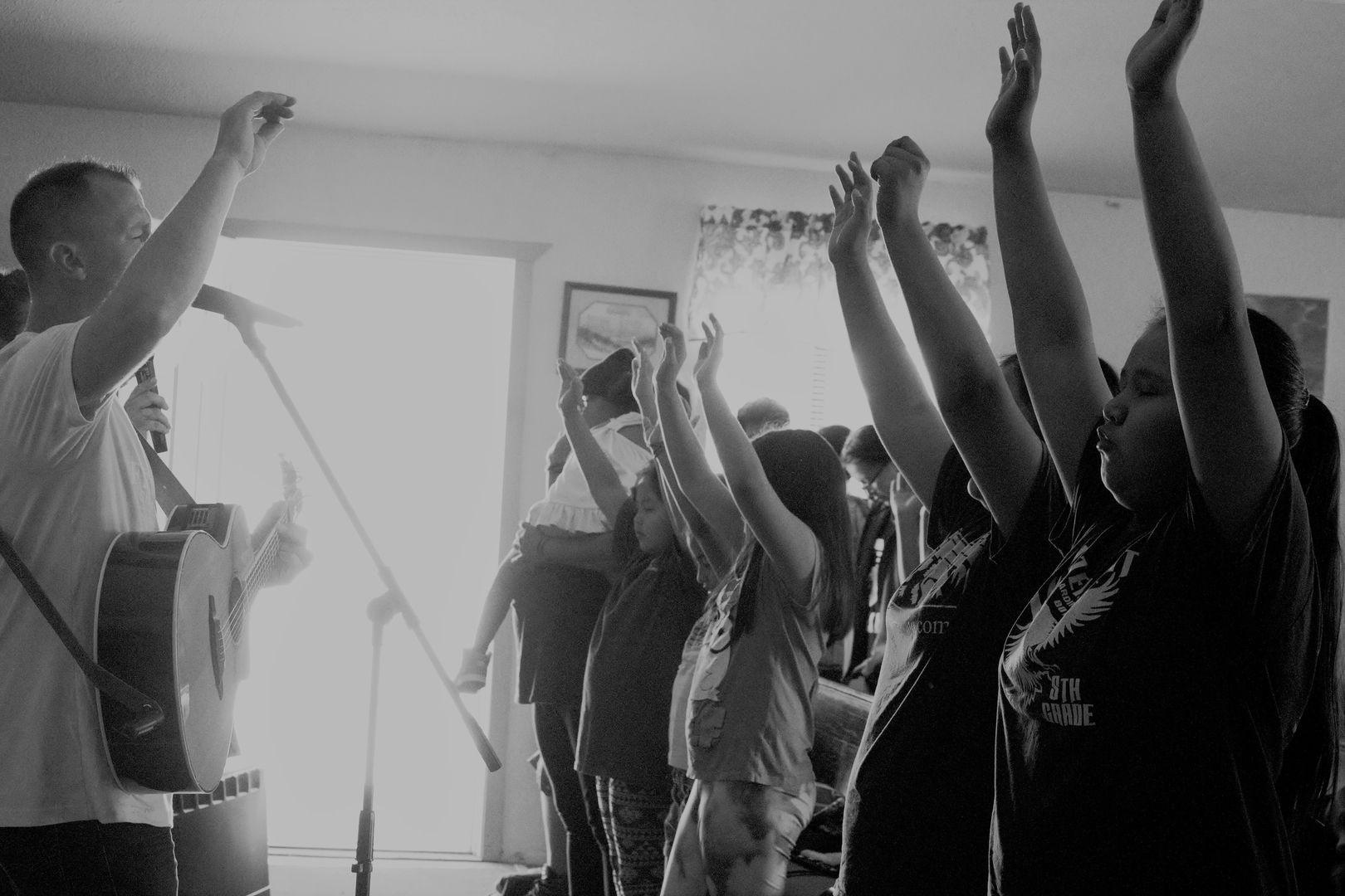 People raising arms in worship, a man with a guitar leading the song indoors.