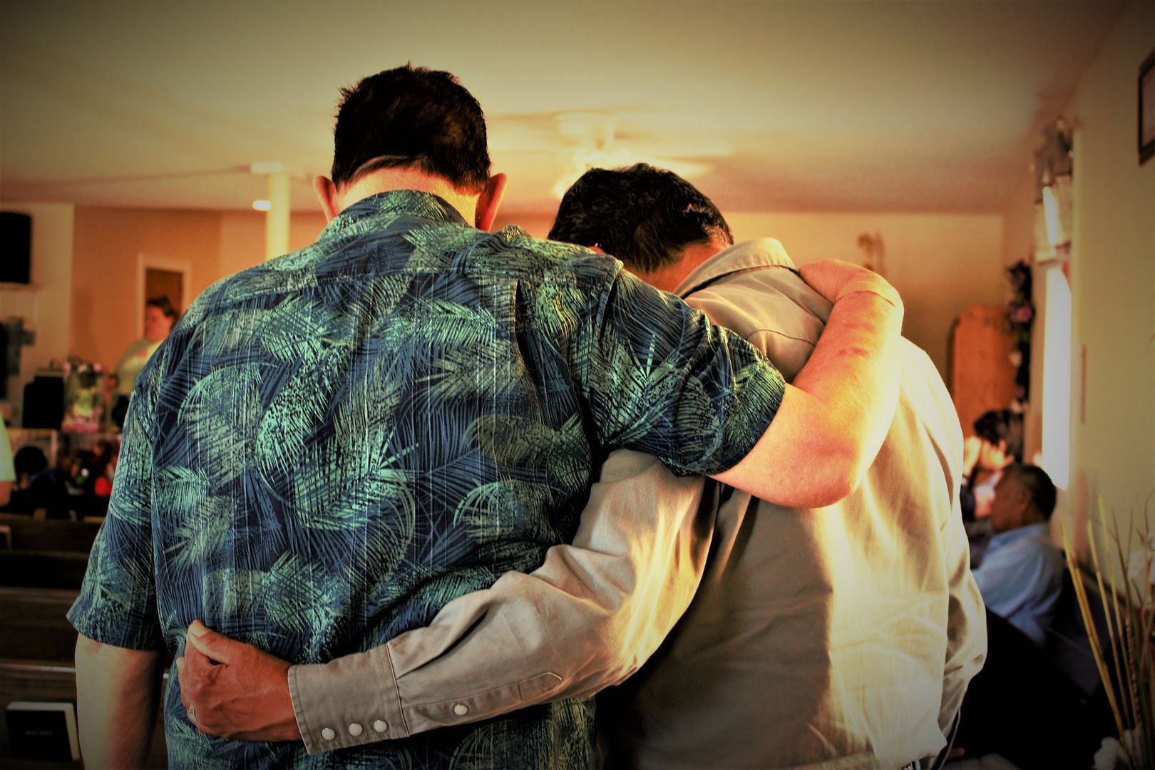 Two men with arms around each other's shoulders; one in a floral shirt, the other in a light shirt, indoors.