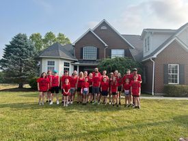 Group of people in red shirts stand in front of a brick house on a grassy lawn.