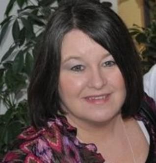Woman with dark hair smiles, wearing a pink and purple patterned top, in front of greenery.