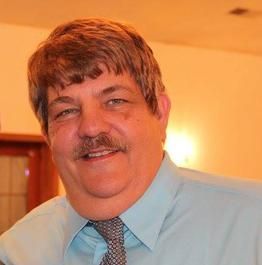 Man with brown hair and mustache wearing a blue shirt and patterned tie, smiling.