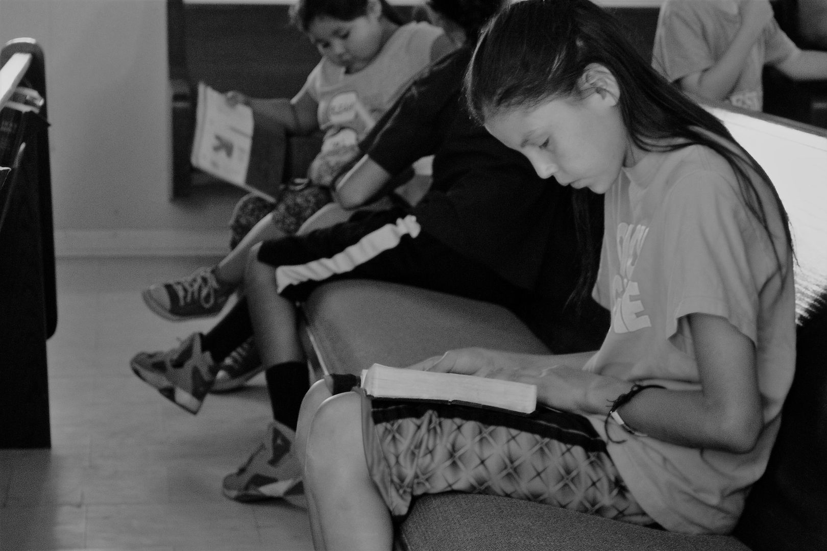 Children sitting in church pews, reading books. Focus on a girl intently reading. Black and white.