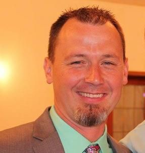 Man in a suit smiles, with a goatee and short brown hair, against an orange background.