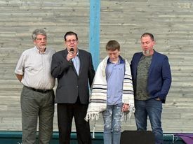 Four men on stage; one speaks into microphone, boy in shawl, backdrop of wood panels.