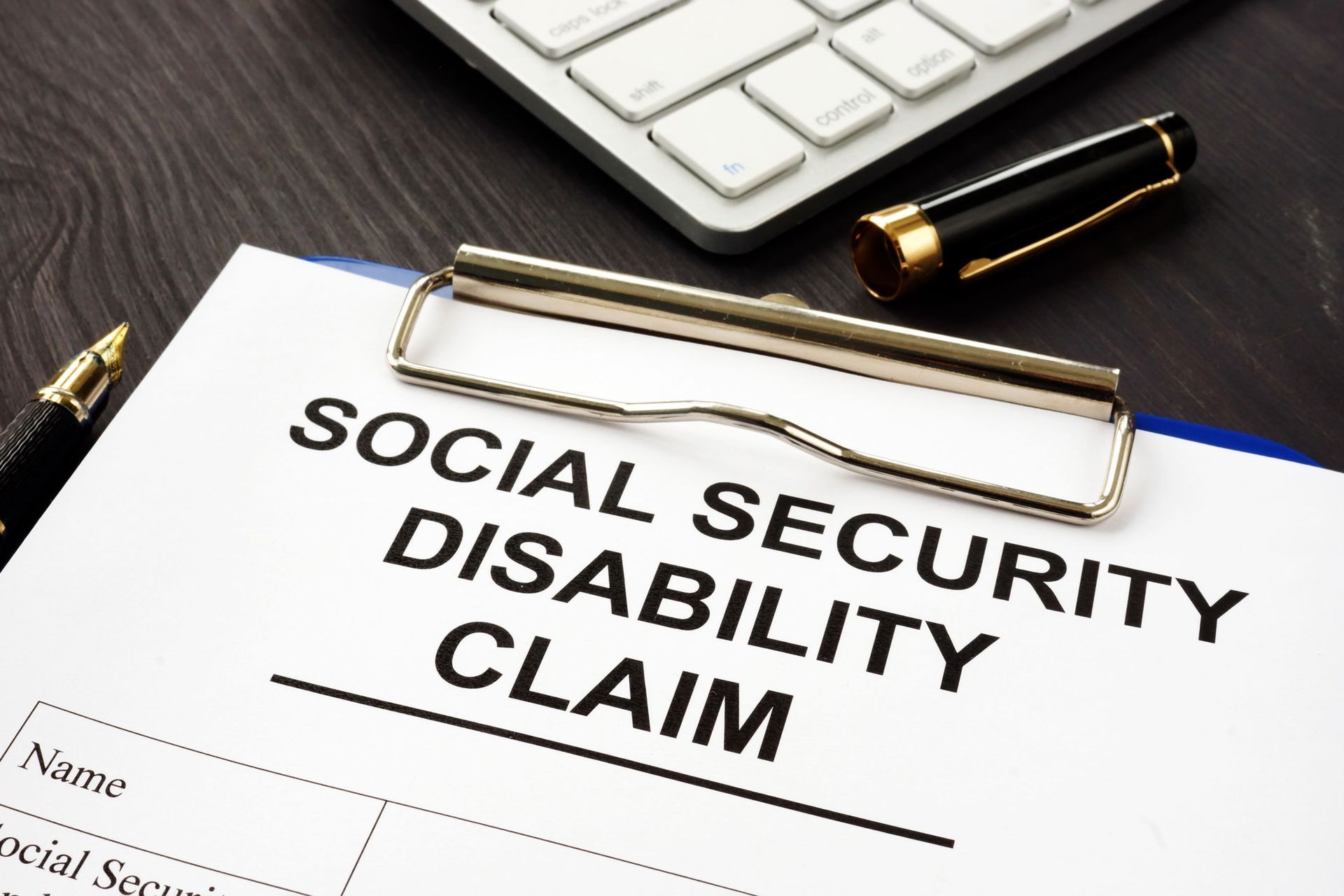 Social Security Disability Claim form on clipboard with keyboard and pen on a wood surface.