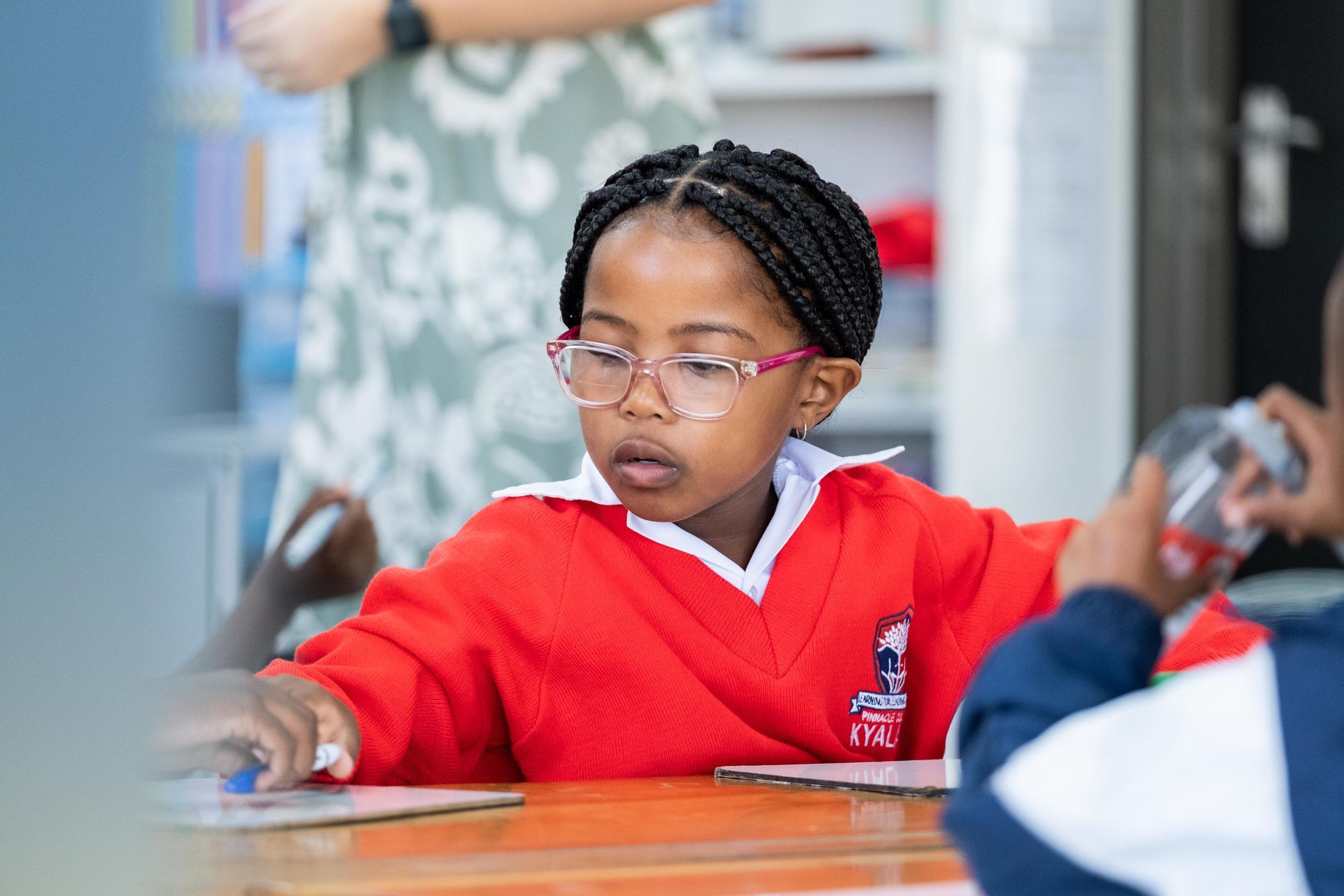 A young girl wearing glasses is sitting at a desk in a classroom.