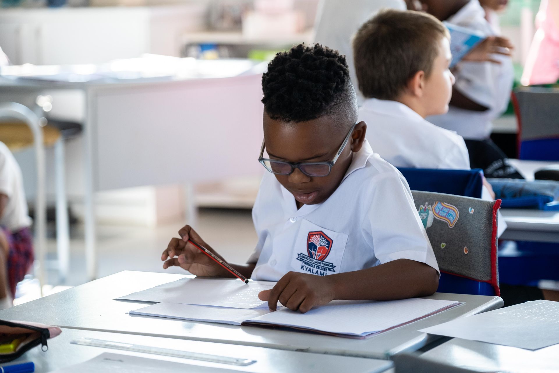 A young boy is sitting at a desk in a classroom writing in a notebook.