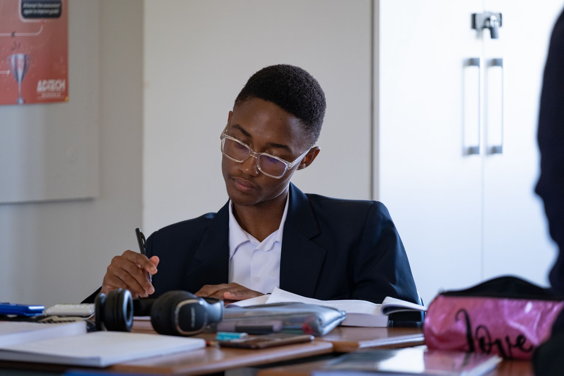 A young man is sitting at a desk in a classroom writing in a notebook.